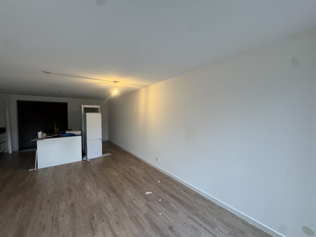 Woonkamer met laminaatvloer en gladde witte wanden, met de keuken op de achtergrond